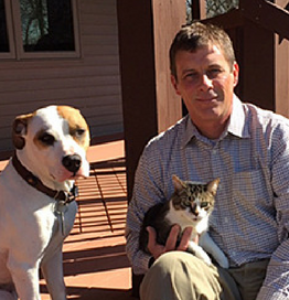 Bio photo of Richard Angelo, Jr. sitting on a porch, holding a kitten as a brown and white dog sits by